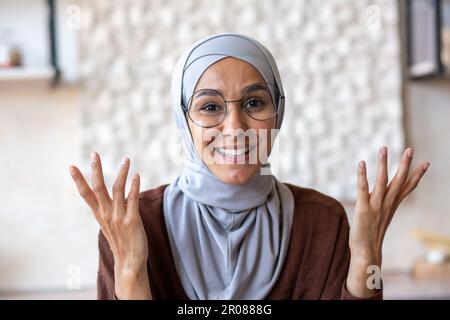 Primo piano. Giovane donna araba in hijab e occhiali che parlano con la macchina fotografica da casa. Gestendo con le mani, sorridendo. Foto Stock