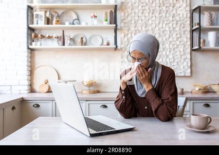 Giovane donna araba in hijab che lavora a casa su un computer portatile in cucina. Non si sente bene, è malata, si asciuga il naso con un tovagliolo da un naso che cola. Foto Stock