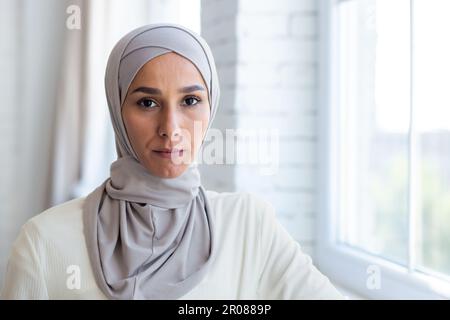 Primo piano. Ritratto di una giovane donna musulmana in un hijab in piedi accanto alla finestra di casa, guardando con fiducia e serietà la macchina fotografica. Foto Stock