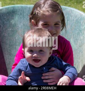 Immagine di due adorabili bambini sorridenti che si tengono all'interno di una carriola. Amore fraterno come giocano in giardino Foto Stock