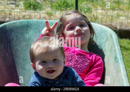 Immagine di due adorabili bambini sorridenti che si tengono all'interno di una carriola. Amore fraterno come giocano nel giardino con la loro sorella fare Foto Stock