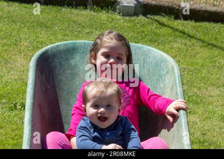 Immagine di due adorabili bambini sorridenti che si tengono all'interno di una carriola. Amore fraterno come giocano in giardino Foto Stock