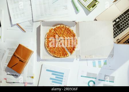 Vista superiore della pizza al tavolo con documenti e computer portatile. Foto Stock