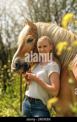 Una giovane donna bionda e il suo cavallo haflinger che si divertono in primavera all'aperto. Scena di amicizia tra una donna equestre e il suo pony Foto Stock