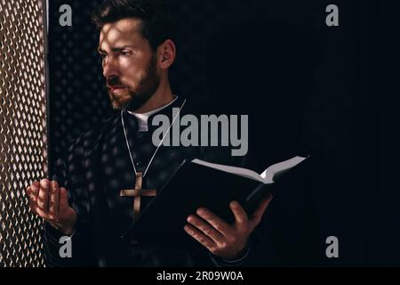 Sacerdote cattolico in cassock tenendo la Bibbia e parlando con parrocchiano in confessionale Foto Stock