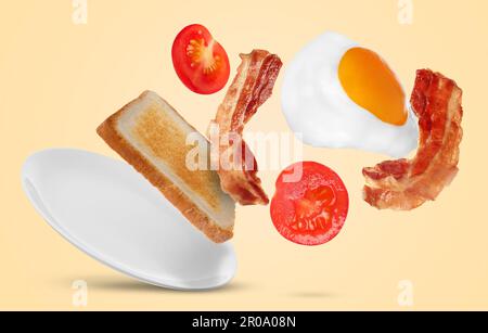 Gustoso uovo di pollo fritto, pancetta, pomodoro e pane tostato che cade nel piatto su sfondo colorato Foto Stock