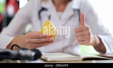 Medico che tiene la pera gialla e che mostra il segno pollice-in su Foto Stock