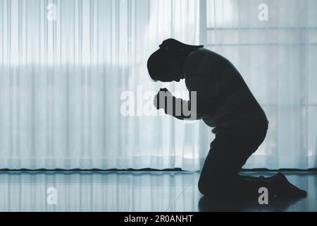 silhouette di donna che si inginocchia e prega nella chiesa moderna al tramonto. Il culto cattolico femminile di preghiera a Dio augura una vita migliore a casa all'alba Foto Stock