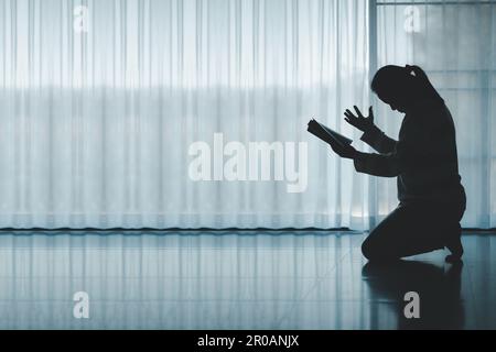 silhouette di donna che si inginocchia e prega nella chiesa moderna al tramonto. Il culto cattolico femminile di preghiera a Dio augura una vita migliore a casa all'alba Foto Stock