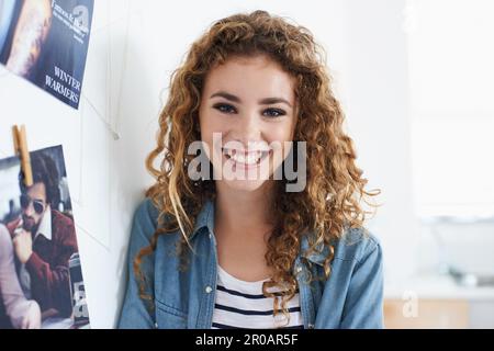 Dopo una carriera nel design grafico. Una giovane grafica sorridente alla fotocamera mentre si trova accanto a una disposizione di foto su una parete. Foto Stock