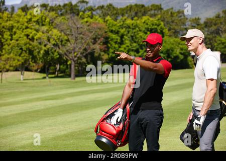 Il foro seguente è quello senso. due uomini che trasportano i loro sacchetti di golf attraverso un campo da golf e che indicano qualcosa. Foto Stock