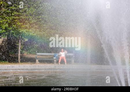 Lady seduto su una panchina nel Parc de Bruxelles, visto attraverso una fontana con effetto arcobaleno. Foto Stock
