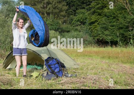 Giovane donna che scuote il sacco a pelo e sorride nella foresta, Baviera, Germania Foto Stock