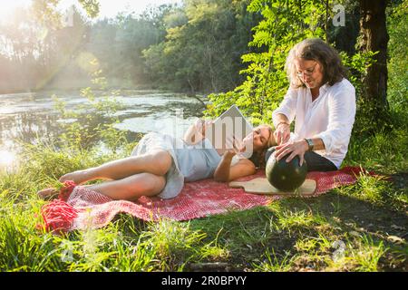 Coppia al pic-nic utilizzando tablet con anguria sul fiume, Baviera, Germania Foto Stock