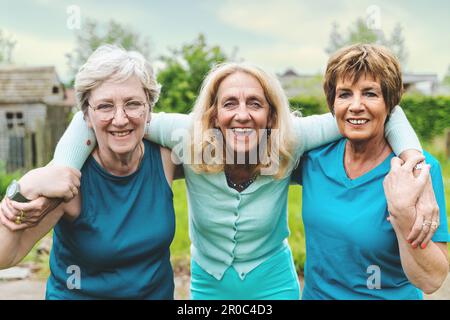 Tre donne nordiche felici negli anni sessanta abbracciano e sorridono alla macchina fotografica dopo un allenamento all'aperto, posandosi in un cortile di campagna. Foto Stock