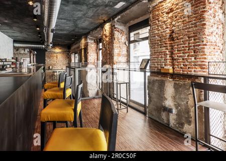 Un banco bar locale con alte poltrone con posti a sedere gialli, pavimenti industriali in parquet e pareti in mattoni a vista e aria condizionata con tubi metallici Foto Stock