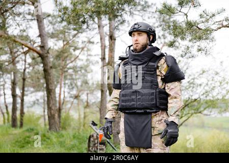 Il giovane soldato in uniformi e gilet tattico lavora nella foresta e si prepara all'azione in una base forestale temporanea. Un uomo fa nel lavoro di sminamento Foto Stock