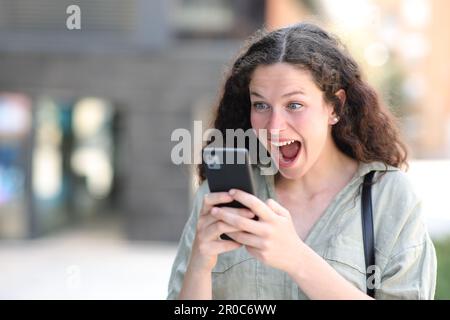 Donna eccitata a guardare contenuti straordinari sul telefono che cammina per strada Foto Stock