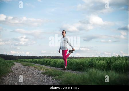 Giovane donna con leggings rosa che si allunga e si scalda prima di andare per una corsa in piedi sulla strada di campagna nel mezzo di bei campi di primavera verde. Foto Stock