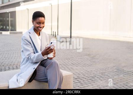 ritratto orizzontale di un'elegante giovane afroamericana. È seduta vicino all'edificio degli uffici e utilizza il suo smartphone Foto Stock