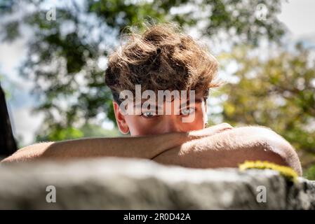 Il giovane cade la testa giù sulle braccia, sorridendo e guardando la telecamera in un parco pubblico Foto Stock