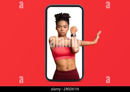 Giovane donna afro-americana slim fiduciosa in abbigliamento sportivo che fa esercizi di stretching per le mani sullo smartphone Foto Stock