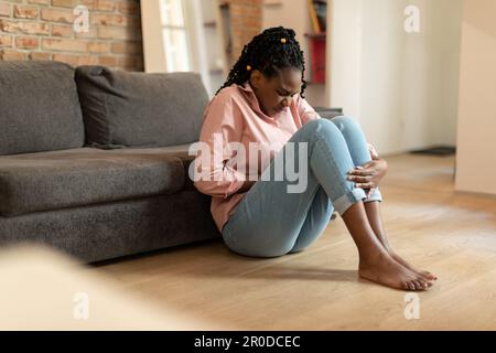 Donna nera che soffre di dolore mestruale, toccando la pancia, avendo crampi addominali durante il periodo, seduta sul pavimento Foto Stock