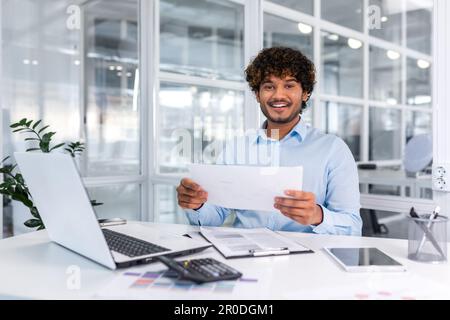 Ritratto di un giovane uomo d'affari ispanico di successo all'interno dell'ufficio, uomo sorridente e guardando la macchina fotografica, lavoratore di carta felice con i risultati di realizzazione seduto sul posto di lavoro con il laptop. Foto Stock