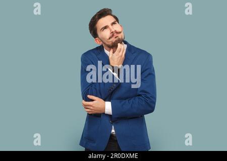 Ritratto di serio uomo d'affari premuroso con baffi in piedi e pensando al suo lavoro, tenendo il mento, indossando il vestito ufficiale di stile. Studio in interni isolato su sfondo azzurro. Foto Stock