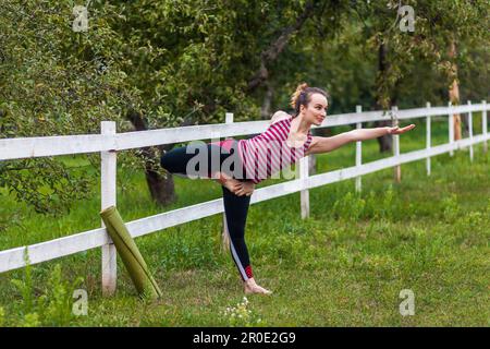 Ritratto a tutta lunghezza di giovane donna sportiva attraente indossare abbigliamento sportivo pratica yoga, facendo esercizio Natarajasana, posa Signore della danza, allenarsi in parco con la bella natura. Foto Stock