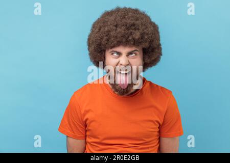 Ritratto dell'uomo con acconciatura afro che indossa una T-shirt arancione che mostra la lingua e gli occhi incrociati con una grimace disobbediente naughty, che fa il viso. Studio in interni isolato su sfondo blu. Foto Stock