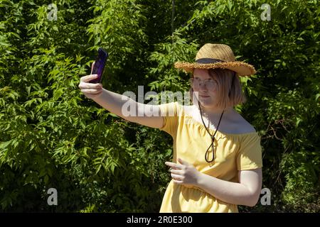 Ragazza asiatica in occhiali, cappello di paglia e vestito giallo spara video sul suo telefono, prende selfie, comunica online, blogger Foto Stock