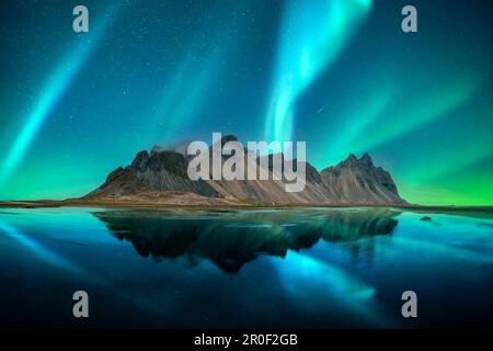 Aurora borealis aurora boreale sulle famose montagne Stokksnes sul capo Vestrahorn. Riflessione nelle acque limpide sullo sfondo dei cieli epici, l'Islanda. Fotografia di paesaggi Foto Stock