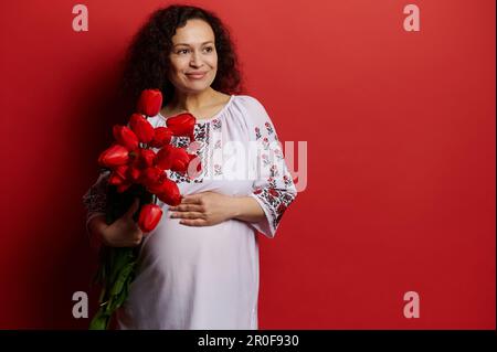 Donna incinta in abito bianco con ornamento ricamato in stile ucraino, carezzando il ventre e posando con tulipani rossi Foto Stock