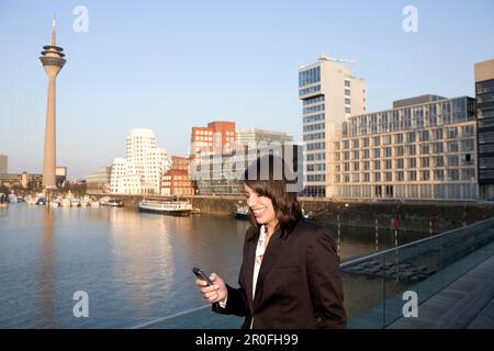 Giovane donna d'affari che scrive un SMS, di fronte allo skyline della città, Media Harbour, Düsseldorf con torre televisiva e Zollhof, architettura di Frank Foto Stock