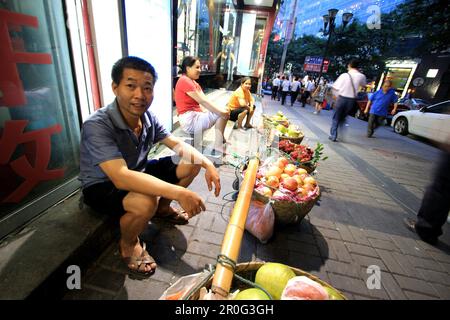 Sorridente falco di strada è seduto di fronte alla sua borsa di frutta a Chongqing, Cina, Asia Foto Stock