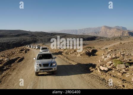 Una fila di veicoli per tutti i terreni che guidano su una strada attraverso paesaggi aridi, montagne al Hajar, Oman, Asia Foto Stock