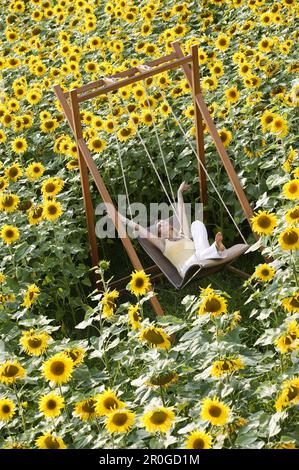 Giovane donna seduta su un'altalena in un campo pieno di girasoli, Baviera, Germania Foto Stock