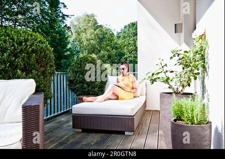 Donna seduta su un lettino mentre usa un iPad su un balcone, Amburgo, Germania Foto Stock