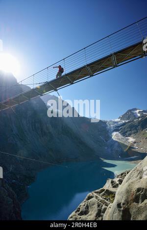 Escursionista a piedi attraverso un ponte sospeso su un lago di montagna, il ghiacciaio Trift sullo sfondo, il ponte sospeso del ghiacciaio Trift, Tieralplistock, Urner Foto Stock