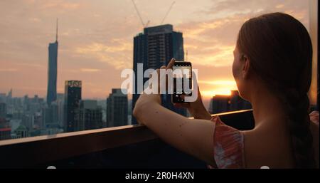 Donna sul tetto di grattacielo spara un paesaggio urbano su un telefono cellulare durante il tramonto. Vista posteriore della ragazza turistica in twiligh in vacanza. Kuala Lumpur Foto Stock