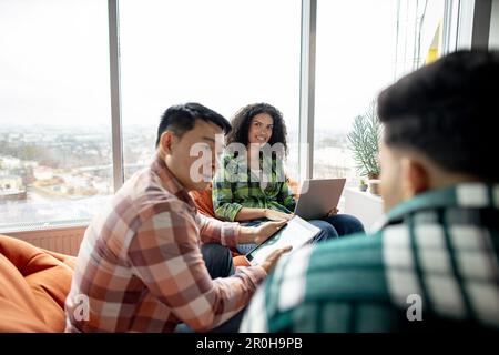 Vista da spalla di allegra donna multirazziale in posa con il notebook mentre i compagni di squadra maschili parlano nello spazio dei colleghi. Diversi professionisti che si scambiano idee sullo sviluppo aziendale in ambienti chiusi. Foto Stock