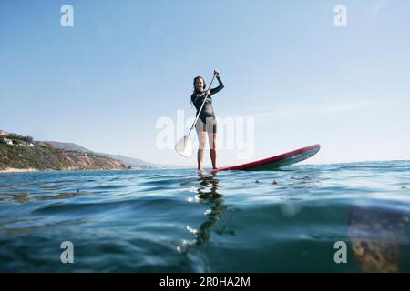 USA, California, Malibu, El Pescador Beach, una donna atletica pagaia la sua tavola da ping-surf lungo la costa di Malibu Foto Stock