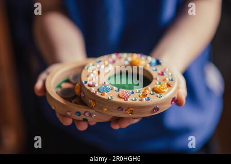 Il bambino risparmia denaro. La banca del piggy dei bambini decorata con i rhinestones nelle mani. Foto Stock