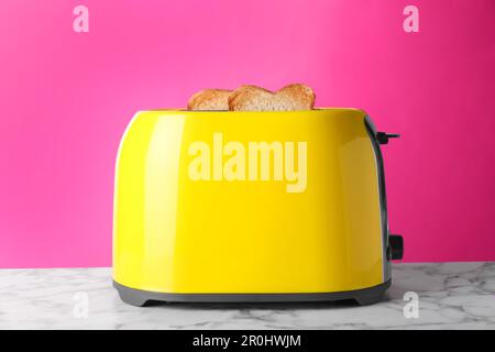 Tostapane giallo con pane tostato su tavolo in marmo bianco su sfondo rosa Foto Stock