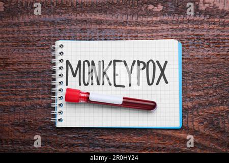 Word Monkeypox scritto in notebook e provetta con campione di sangue su tavolo di legno, vista dall'alto Foto Stock