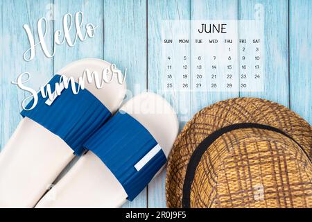 Ciao estate 2023 giugno calendario e slipper di gomma blu con cappello di paglia marrone su sfondo blu. Foto Stock