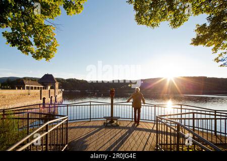 Uomo in piedi su una piattaforma di osservazione alla luce del sole al tramonto, guardando alla diga di Edertalsperre sul Lago Edersee in Kellerwald-Edersee National Pa Foto Stock