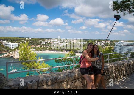 Due giovani donne scattano foto selfie che si affacciano sulla baia di Cala Galdana e sulla spiaggia con smartphone sulla prolunga, Cala Galdana, Menorca, Balearic Isla Foto Stock