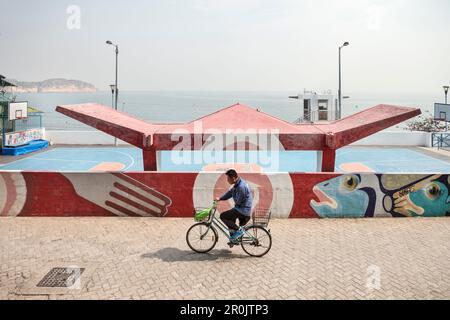 Uomo cinese in bicicletta passa campo da gioco di fronte al mare, Cheng Chau Island, Hongkong, Cina, Asia Foto Stock